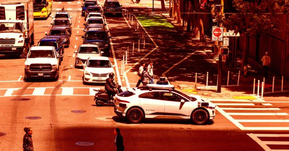 A busy urban street scene with multiple cars lined up in traffic, including a white Waymo self-driving car equipped with sensors on the roof and sides. A person on a scooter is next to the Waymo car, and three pedestrians are crossing the street near the vehicle. There are traffic signs indicating "Do Not Enter" and bicycle signals. Trees line the sidewalk, casting shadows on the street. The overall lighting has a warm, reddish tone.