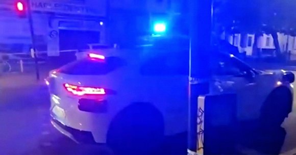 White police car with flashing blue lights at night on a street corner, near a traffic light showing red. A person is standing on the sidewalk to the right of the car.