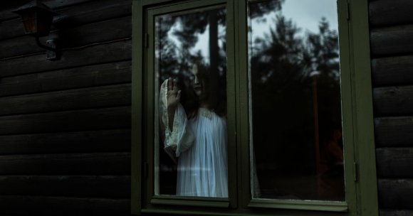 A person with long dark hair and pale face paint, wearing a white flowing garment, stands behind a green-framed window of a dark wooden cabin. Their hand is pressed against the glass, and the reflection of trees is visible on the window. The scene has a dark, eerie atmosphere.