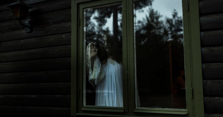 A person with long dark hair and pale face paint, wearing a white flowing garment, stands behind a green-framed window of a dark wooden cabin. Their hand is pressed against the glass, and the reflection of trees is visible on the window. The scene has a dark, eerie atmosphere.