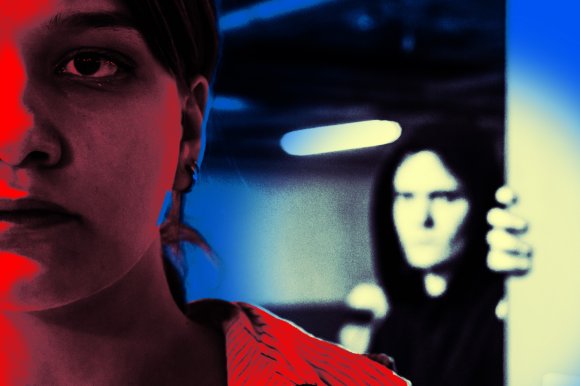 Close-up of a person's face on the left side, illuminated with red light, showing a serious expression. In the background on the right, a blurred figure wearing a dark hooded garment is partially visible, holding onto a vertical surface, with a blue-toned environment behind them.