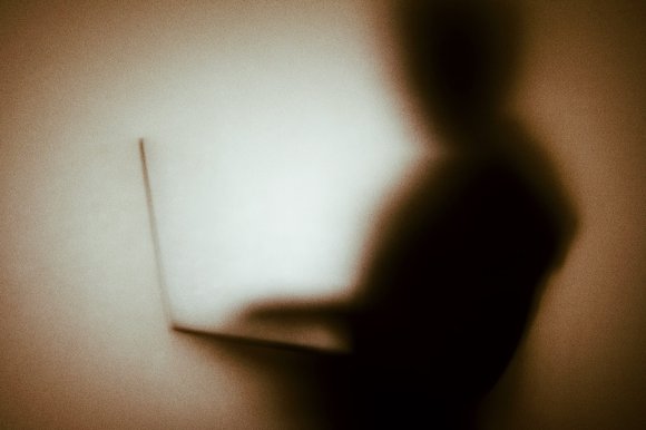 Silhouette of a person using a laptop, with the figure and device appearing as a dark shadow against a softly lit, neutral background. The image has a grainy texture and a warm, muted tone.