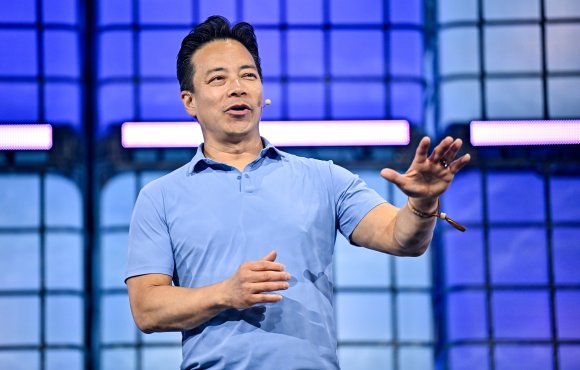 A man wearing a light blue polo shirt is speaking on stage with a microphone headset. He is gesturing with his right hand extended forward and his left hand partially raised. The background features a blue and purple grid-like pattern with illuminated horizontal light bars.