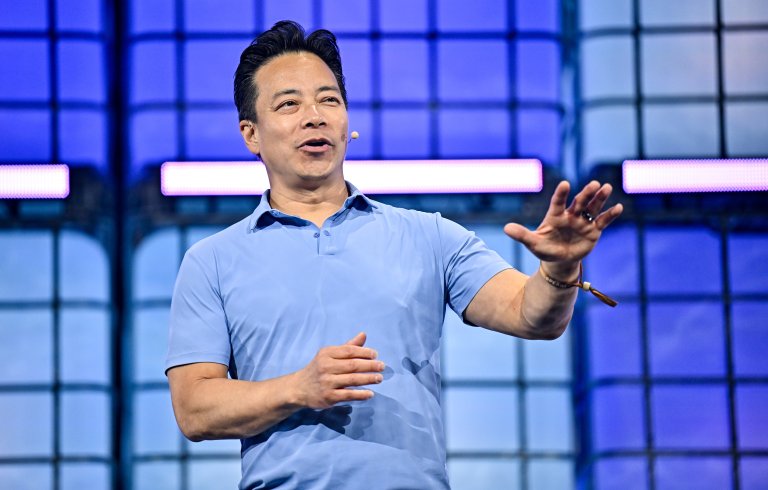 A man wearing a light blue polo shirt is speaking on stage with a microphone headset. He is gesturing with his right hand extended forward and his left hand partially raised. The background features a blue and purple grid-like pattern with illuminated horizontal light bars.