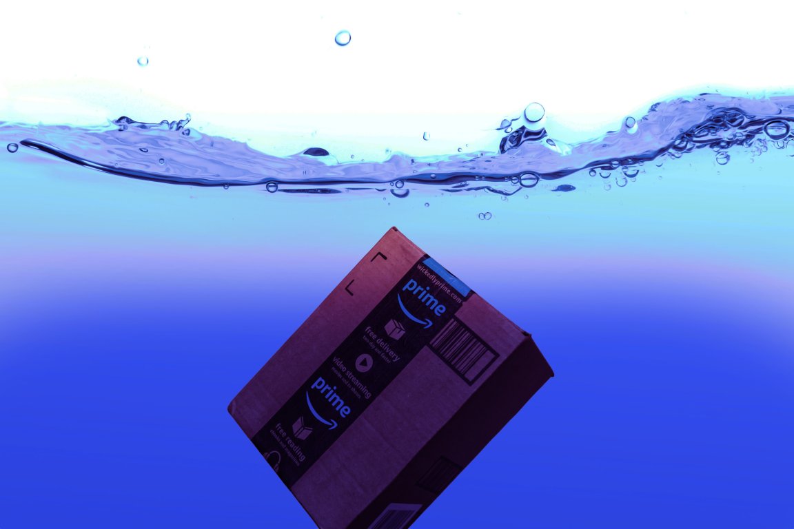 A cardboard Amazon Prime package submerged underwater with visible air bubbles and a clear water surface above. The water has a gradient of blue shades.
