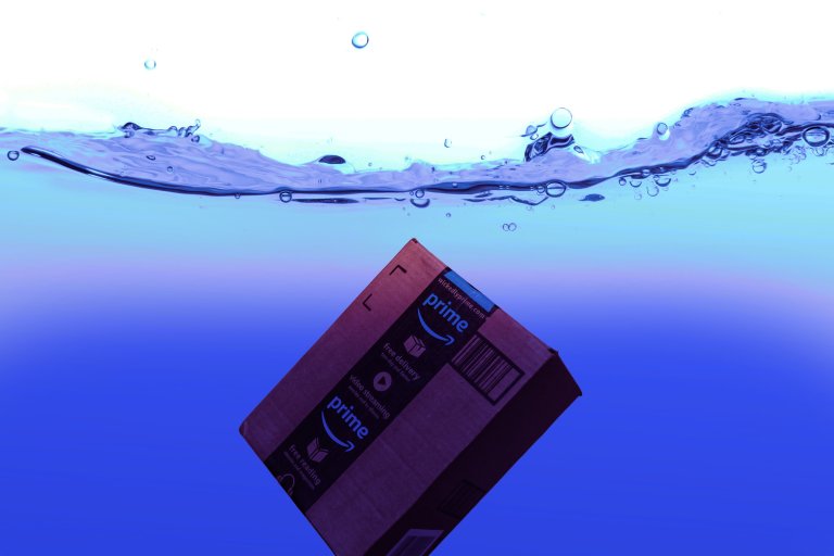 A cardboard Amazon Prime package submerged underwater with visible air bubbles and a clear water surface above. The water has a gradient of blue shades.