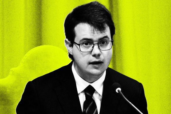 A young man with dark hair and glasses is speaking into a microphone. He is wearing a black suit, white shirt, and striped tie. The background is bright yellow with a blurred chair visible behind him. The image has a high-contrast, stylized effect.