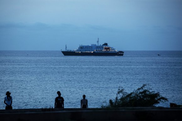 A large, multi-deck ship with lights on is sailing on a calm sea during dusk or early evening. In the foreground, three silhouetted people stand near the shore, with some vegetation visible on the right side. The sky is mostly clear with a soft blue hue.