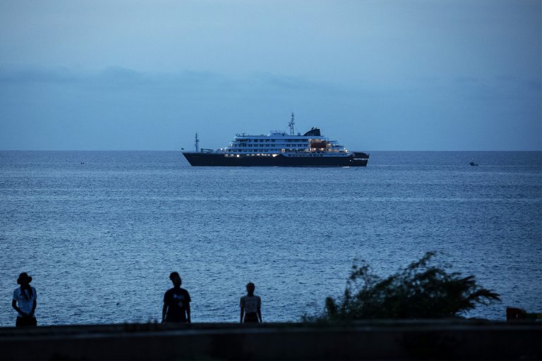 A large, multi-deck ship with lights on is sailing on a calm sea during dusk or early evening. In the foreground, three silhouetted people stand near the shore, with some vegetation visible on the right side. The sky is mostly clear with a soft blue hue.