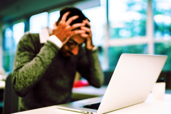 A person sitting at a desk with a laptop, holding their head in their hands, appearing stressed or frustrated. The background shows large windows with natural light coming in.