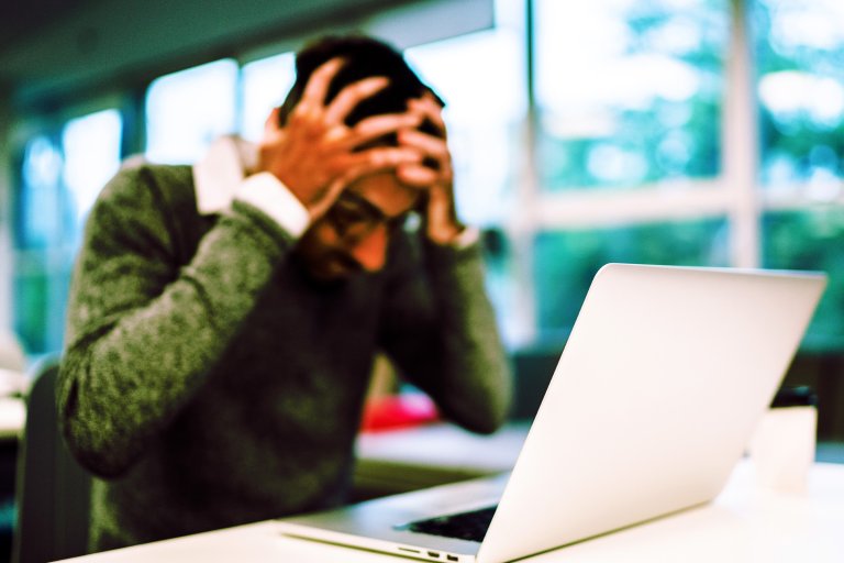 A person sitting at a desk with a laptop, holding their head in their hands, appearing stressed or frustrated. The background shows large windows with natural light coming in.