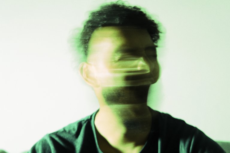 A portrait of a person with short curly hair wearing a dark shirt, captured with motion blur that distorts the face horizontally, creating a ghostly, stretched effect. The background is plain and light-colored.