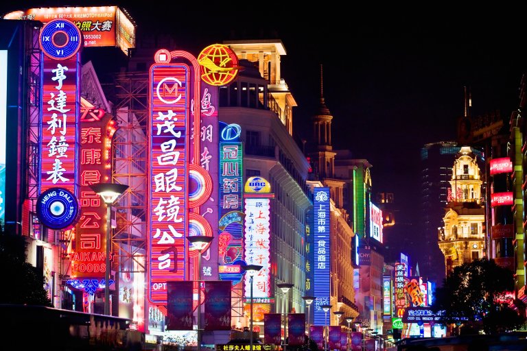 Brightly lit neon signs with Chinese characters line a bustling city street at night, featuring a mix of colorful advertisements in red, blue, yellow, and green.