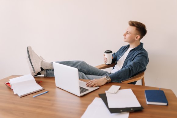 A young man is sitting at a wooden desk with his feet up, wearing white sneakers, light blue jeans, a white t-shirt, and a blue button-up shirt. He is holding a disposable coffee cup in one hand and resting the other hand on a silver laptop. The desk has scattered papers, a blue pen, an open notebook, a closed blue notebook, and a white smartphone. The background is plain and light-colored.