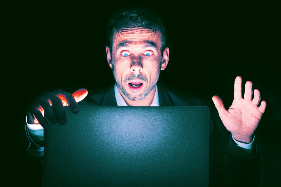 An image of a businessman in the dark opening his laptop up, lit from underneath with one hand held up in shock. He is surprised by what he sees onscreen.