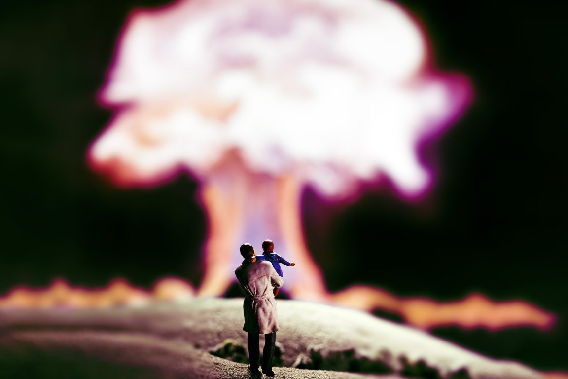 A miniature figure of a person holding a child, both facing away, looking towards a large, blurred mushroom cloud explosion in the background. The scene is dark with the bright explosion providing contrast.