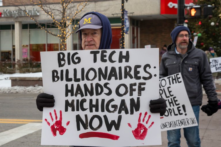 A person wearing a blue hoodie and a gray cap with a yellow "M" holds a protest sign that reads "BIG TECH BILLIONAIRES: HANDS OFF MICHIGAN NOW" with two red handprints and a red underline. Another person in a gray jacket and blue beanie stands in the background holding a partially visible sign. The scene appears to be outdoors in a city with some snow on the ground.