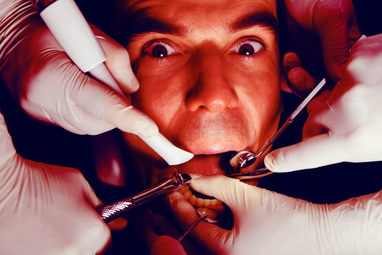 Close-up of a man with wide eyes undergoing dental treatment, surrounded by multiple gloved hands holding various dental instruments inside his open mouth.
