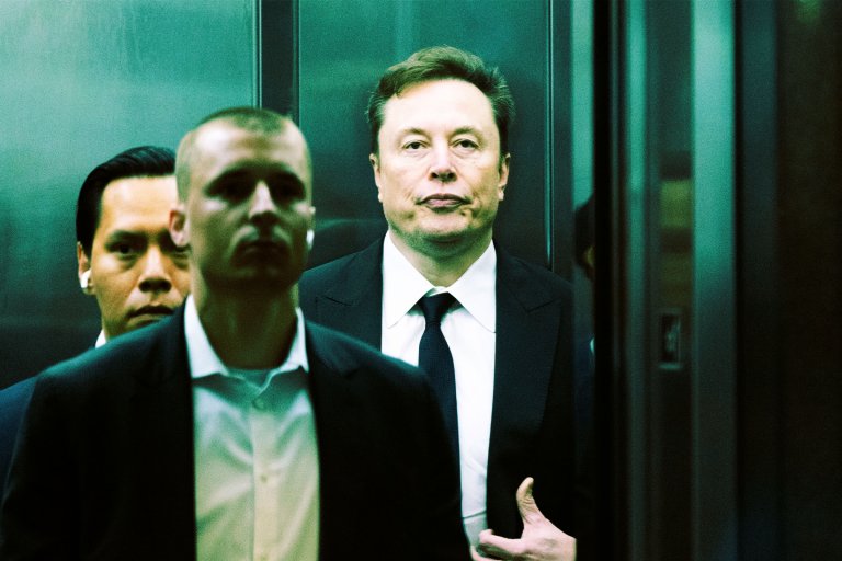 Elon Musk is standing in an elevator, wearing a black suit, white shirt, and black tie.
