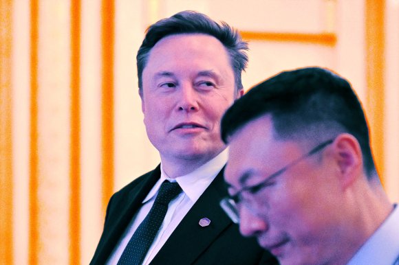 Elon Musk in a black suit, white shirt, and black tie, looking to his left. Another man with glasses and short black hair is in the foreground, slightly out of focus. The background features a warm, softly lit setting with vertical orange and cream-colored stripes.