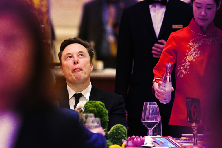 Elon Musk is seated at a formal dining table, wearing a black suit and tie, looking upward with a contemplative expression.