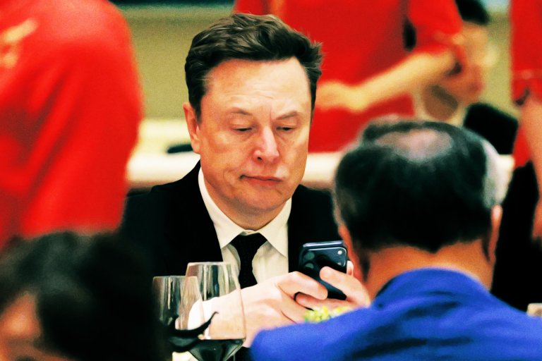 Elon Musk wearing a black suit and tie, sitting at a table and looking at his smartphone. There are wine glasses on the table, and blurred people in red clothing in the background.