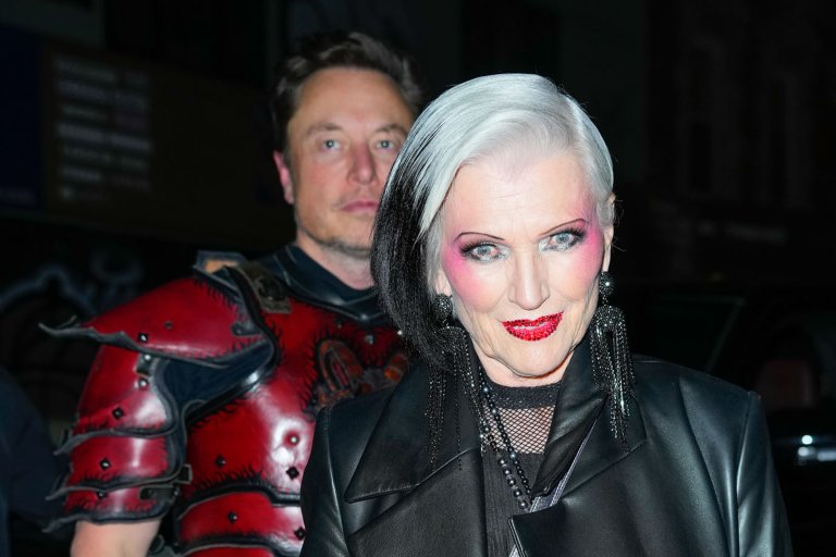Maye Musk is shown with striking black and white hair, bold pink eyeshadow, and glittery red lipstick wears large black earrings and a black leather jacket. Behind her stands Elon Musk in a red and black medieval-style armor costume. The background is dark and out of focus.