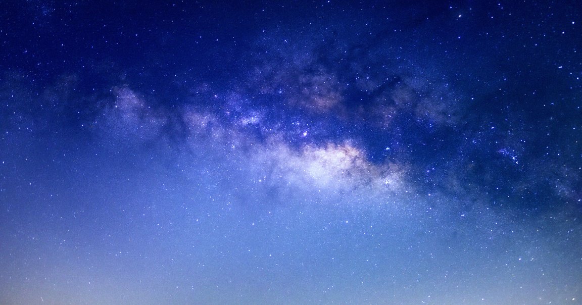 A clear night sky filled with numerous stars and a visible section of the Milky Way galaxy, showing a dense cluster of stars and cosmic dust with a gradient from deep blue at the top to a lighter blue near the horizon.