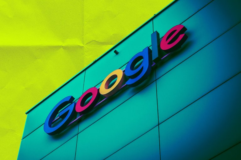 Google logo sign mounted on a teal-colored building facade with a bright yellow background. The letters are in the classic Google colors: blue, red, yellow, blue, and red.