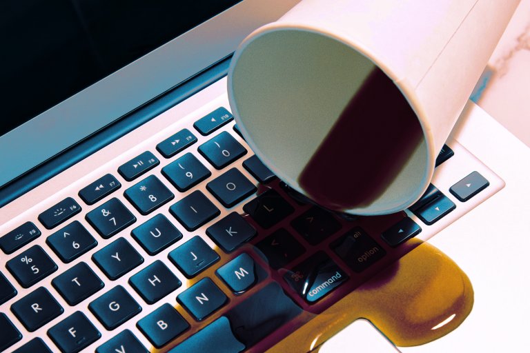 A close-up of a laptop keyboard with a white paper cup tipped over, spilling a dark liquid onto the keys and surrounding area. The liquid is pooling and spreading across the keyboard and laptop surface.