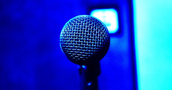 Close-up of a microphone head with a metal mesh grille, illuminated by blue lighting against a blurred blue background.