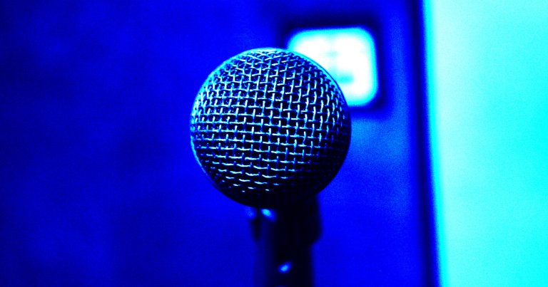 Close-up of a microphone head with a metal mesh grille, illuminated by blue lighting against a blurred blue background.