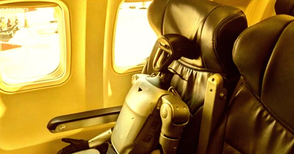 A humanoid robot is seated in an airplane window seat with black leather upholstery. The robot has a sleek, metallic body with a black head featuring a visor-like face. The airplane window shows a bright, sunlit exterior.