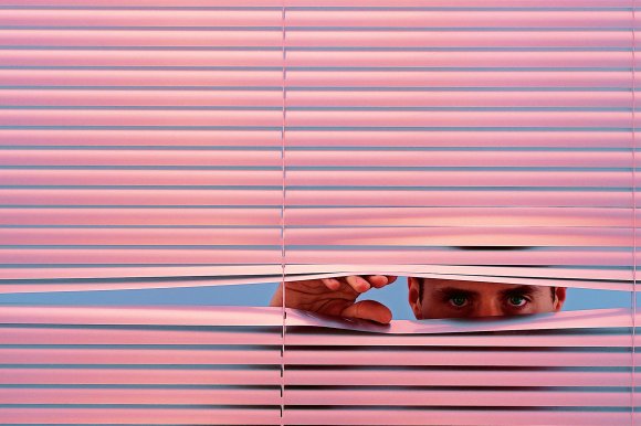 A person with green eyes is peeking through partially opened pink window blinds, using their hand to lift one of the slats. The background behind the blinds is a solid light blue.