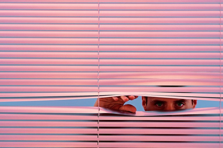 A person with green eyes is peeking through partially opened pink window blinds, using their hand to lift one of the slats. The background behind the blinds is a solid light blue.