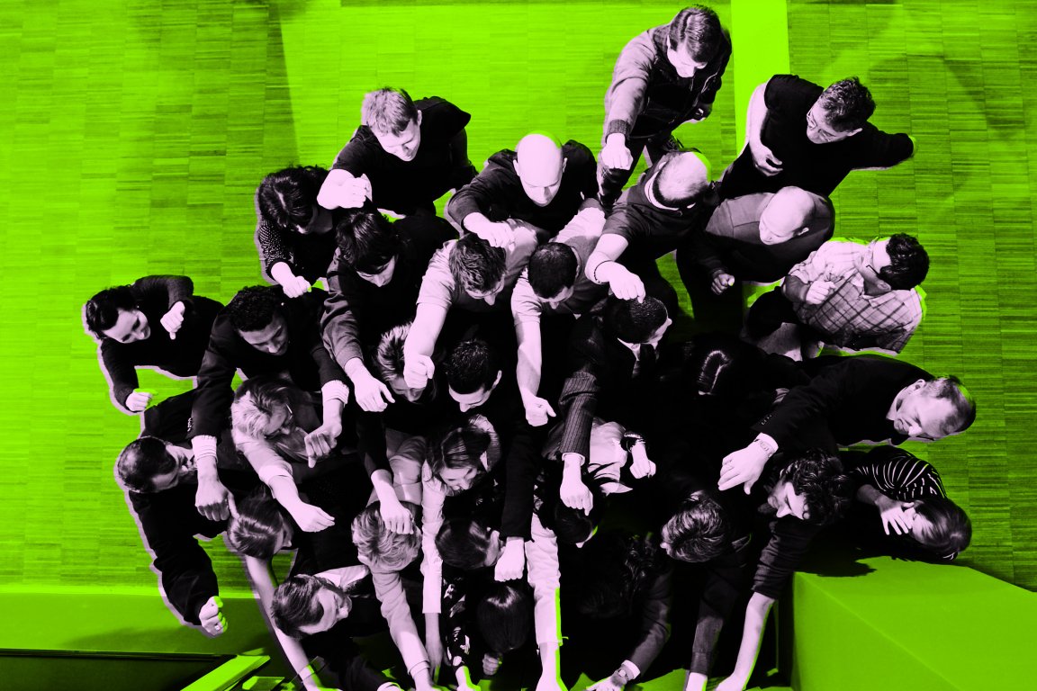 A large group of people standing closely together, viewed from above, all extending one arm forward with a clenched fist. The image has a high-contrast effect with the people in black and white and the floor in bright green. The individuals appear engaged in a collective or unified gesture.