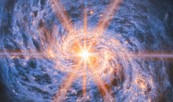 A bright, glowing object at the center of a swirling blue and orange spiral galaxy, with radiant light beams extending outward in multiple directions. The galaxy's spiral arms are detailed with clusters of stars and cosmic dust.