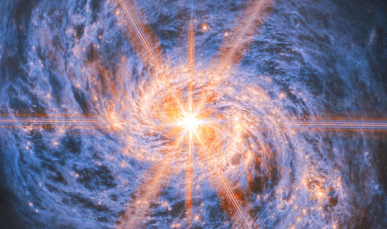 A bright, glowing object at the center of a swirling blue and orange spiral galaxy, with radiant light beams extending outward in multiple directions. The galaxy's spiral arms are detailed with clusters of stars and cosmic dust.