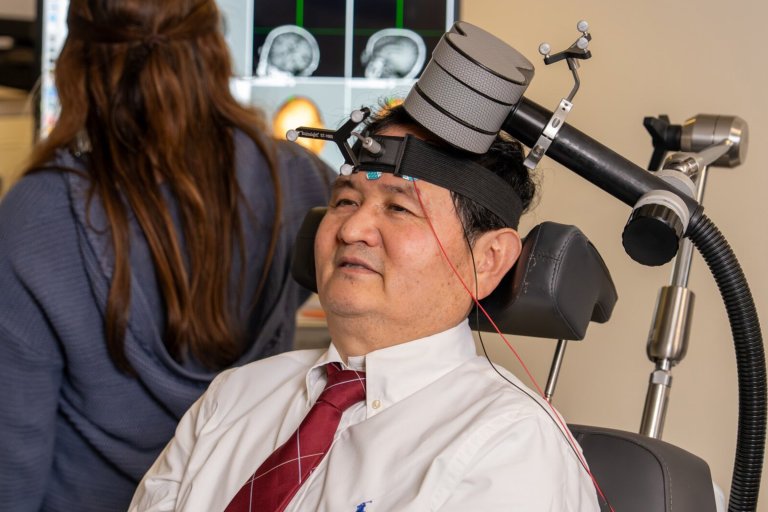 A man wearing a white shirt and red tie is seated with a head-mounted device attached to his forehead, connected to a large mechanical arm. In the background, a woman with long brown hair, wearing a blue sweater, is facing away from the camera. A computer screen displaying brain scans is visible behind them.
