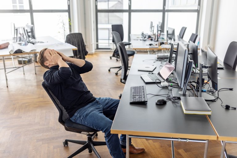 A man sitting in an office chair at a desk with multiple computer monitors, laptops, a keyboard, and a mouse. He is leaning back with his hands covering his face, appearing stressed or frustrated. The office has large windows and wooden flooring, with several empty chairs and desks in the background.