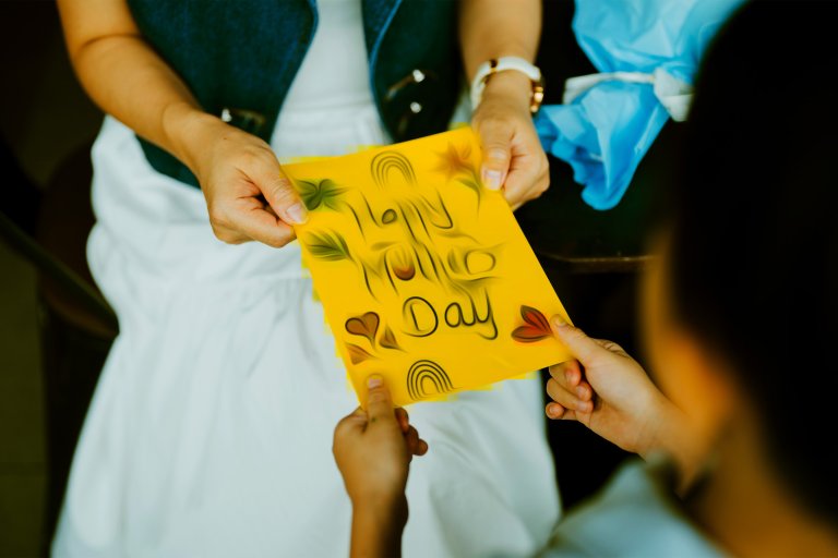 Two people are holding a yellow card with the words "Happy Mother's Day" written on it, but distorted as though it was an AI generation.