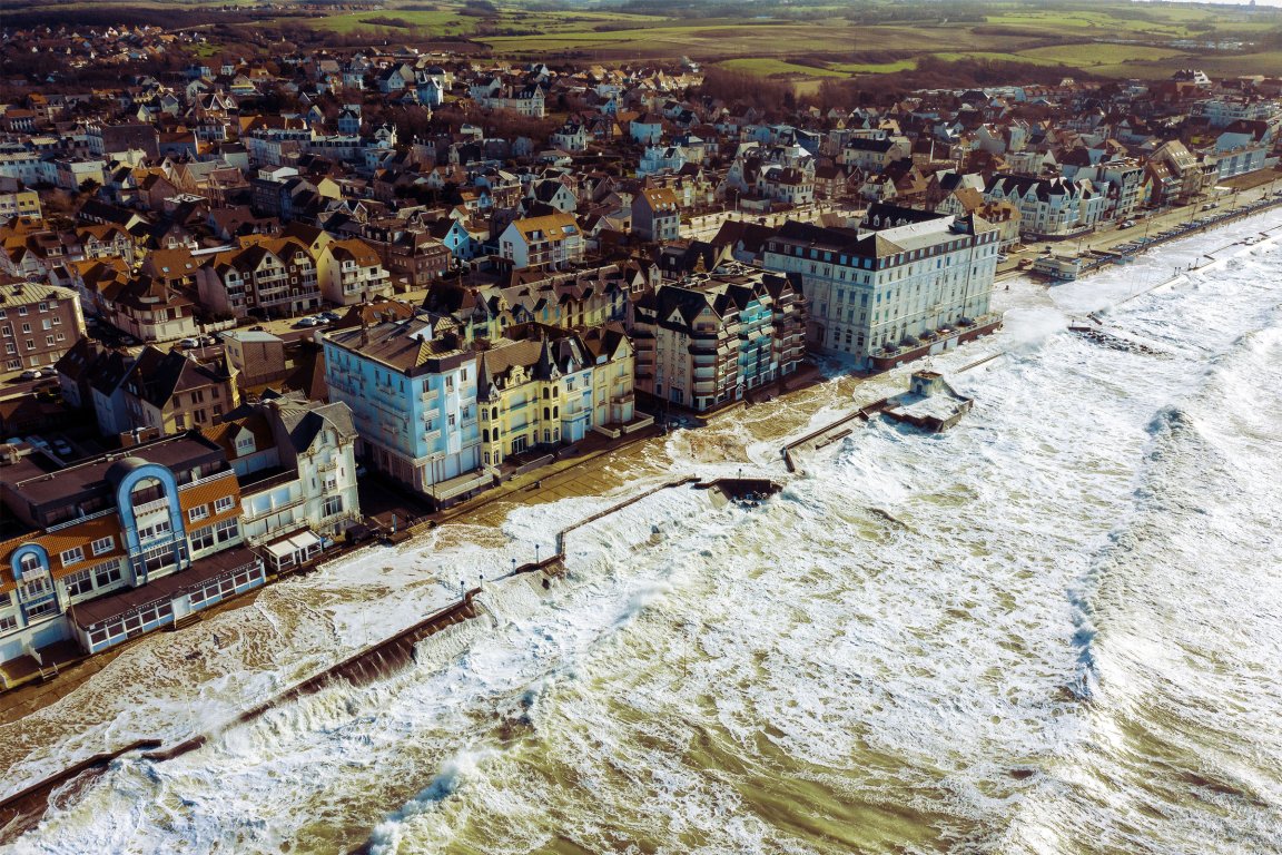 A coastal town with a densely packed cluster of buildings along the shoreline. The buildings are mostly multi-story with varied architectural styles, including some with pointed roofs and balconies. The sea is rough, with large waves crashing against the seawall and flooding the promenade. Beyond the town, there are green fields and open land. The overall scene suggests a windy, possibly stormy day by the coast.