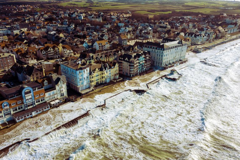 A coastal town with a densely packed cluster of buildings along the shoreline. The buildings are mostly multi-story with varied architectural styles, including some with pointed roofs and balconies. The sea is rough, with large waves crashing against the seawall and flooding the promenade. Beyond the town, there are green fields and open land. The overall scene suggests a windy, possibly stormy day by the coast.