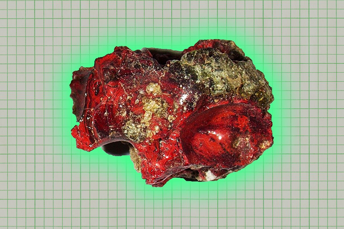 A rough, irregularly shaped mineral or rock specimen with a glossy surface, predominantly red with patches of greenish and yellowish areas. The background is a grid pattern.