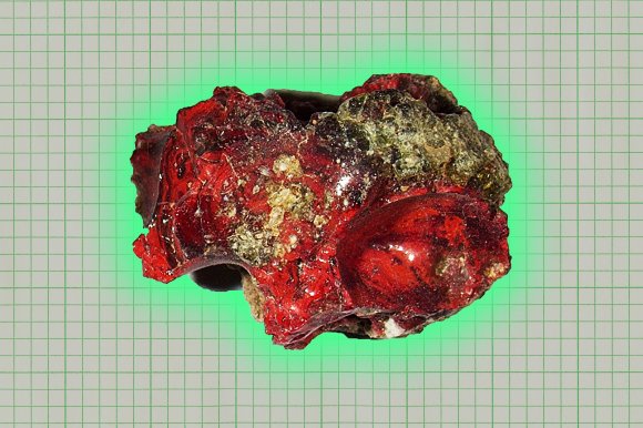 A rough, irregularly shaped mineral or rock specimen with a glossy surface, predominantly red with patches of greenish and yellowish areas. The background is a grid pattern.