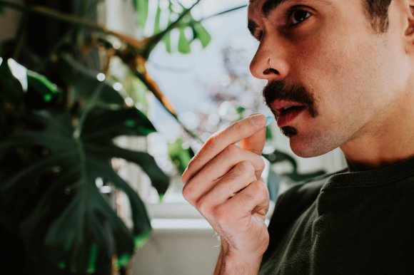 A close-up of a man with a mustache and nose ring holding a small blue pill near his mouth, about to take it. The background includes blurred green leaves and a window with natural light.