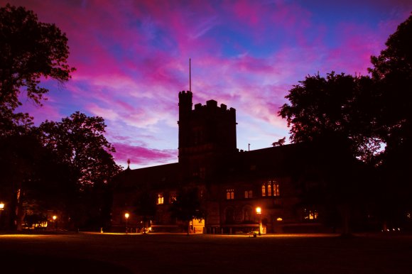 A large, dark silhouette of a historic building at Princeton with a central tower, illuminated from within by warm yellow lights. The building is surrounded by tall trees, and the sky above is a vibrant mix of purple, pink, and blue hues, suggesting either dawn or dusk. Street lamps near the building cast a soft glow on the ground.