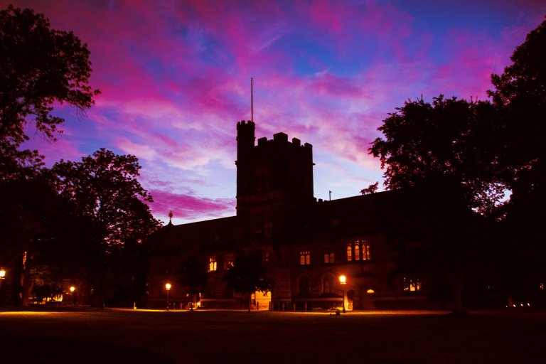 A large, dark silhouette of a historic building at Princeton with a central tower, illuminated from within by warm yellow lights. The building is surrounded by tall trees, and the sky above is a vibrant mix of purple, pink, and blue hues, suggesting either dawn or dusk. Street lamps near the building cast a soft glow on the ground.