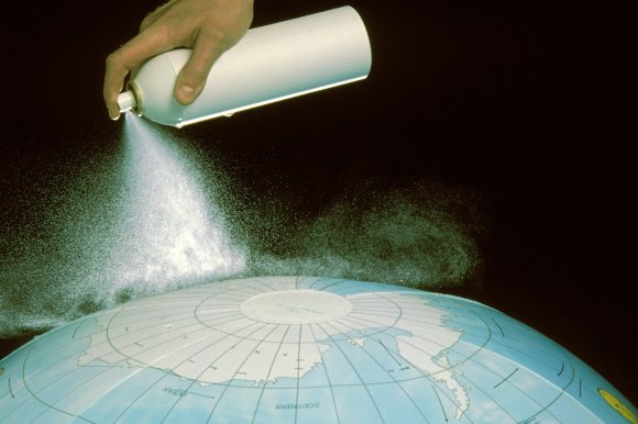 A hand is spraying a white aerosol can over a globe, focusing on the polar region. The spray creates a misty cloud above the globe, which is set against a black background. The globe shows the southern polar area with labeled continents and oceans.