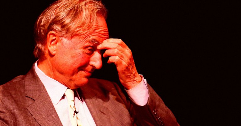 Richard Dawkins wearing a brown suit, white shirt, and light-colored tie, touching his forehead with his right hand against a dark background.
